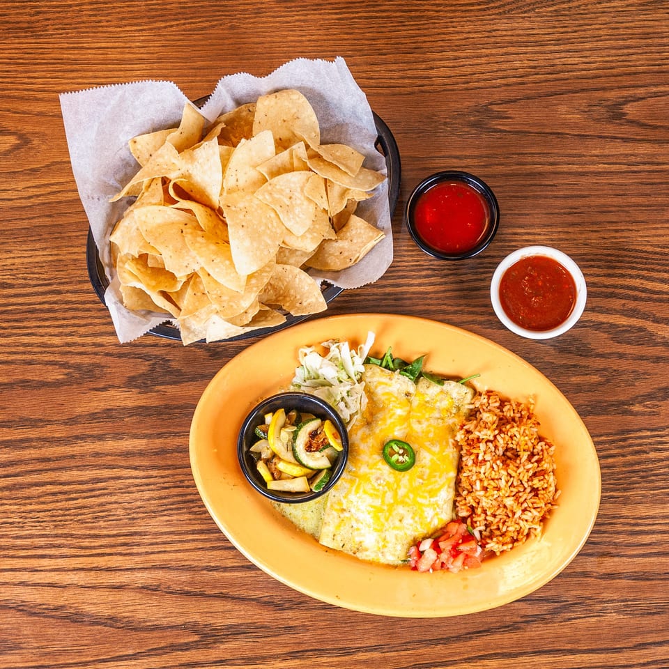 Cheesy enchiladas, Mexican rice, sautéed vegetables, tortilla chips, and salsa on a wooden table.
