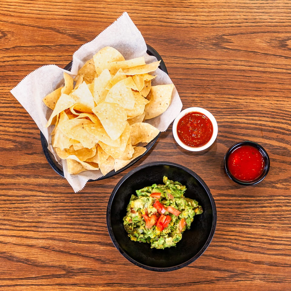Overhead view of tortilla chips, guacamole with tomatoes, and salsa on a wooden table.