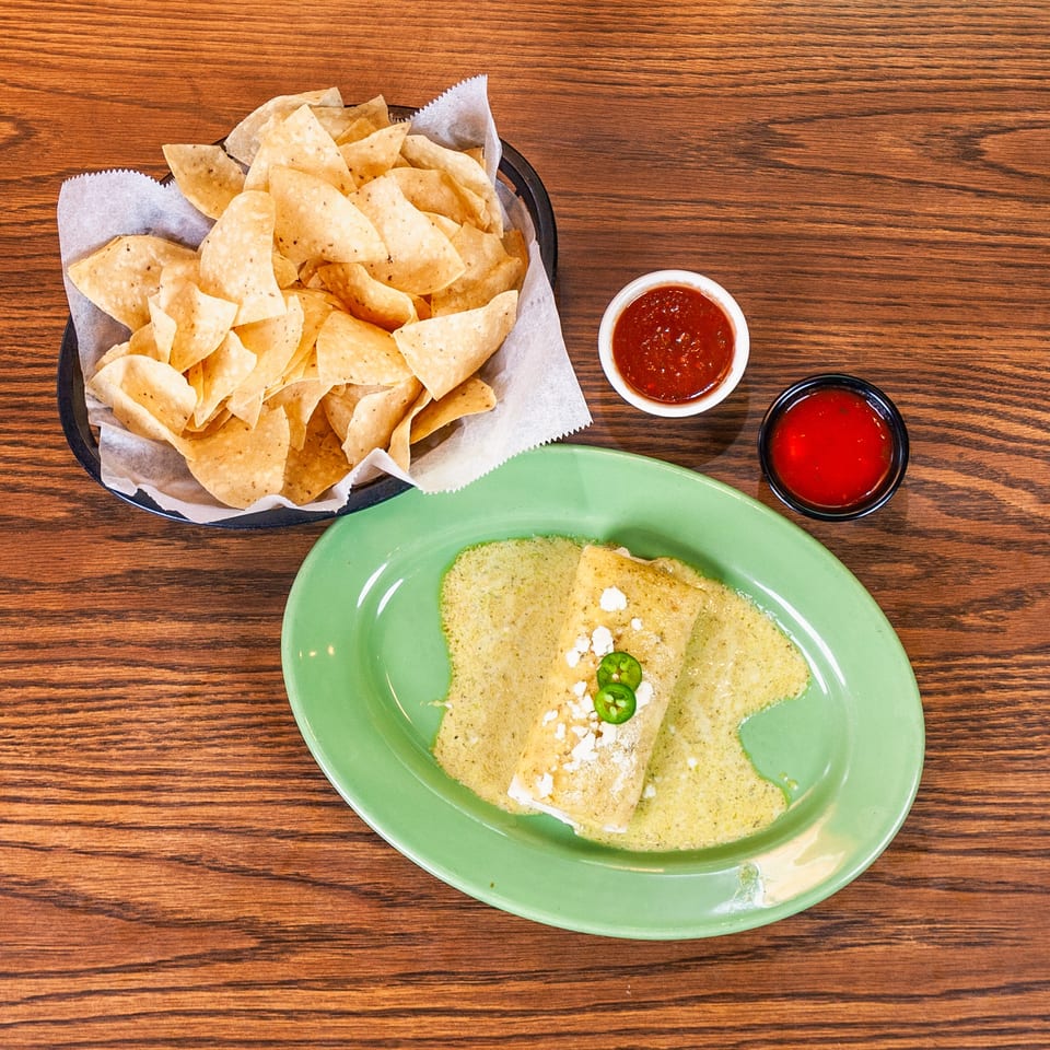 Tortilla chips, a green-sauced burrito with cheese and jalapeños, and two salsa bowls on a wooden table.