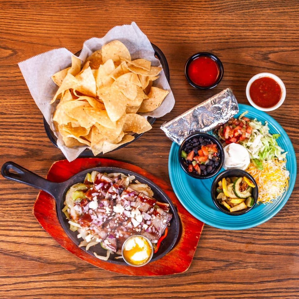 Sizzling fajita platter, blue plate with rice and sides, and a basket of tortilla chips on a wooden table.