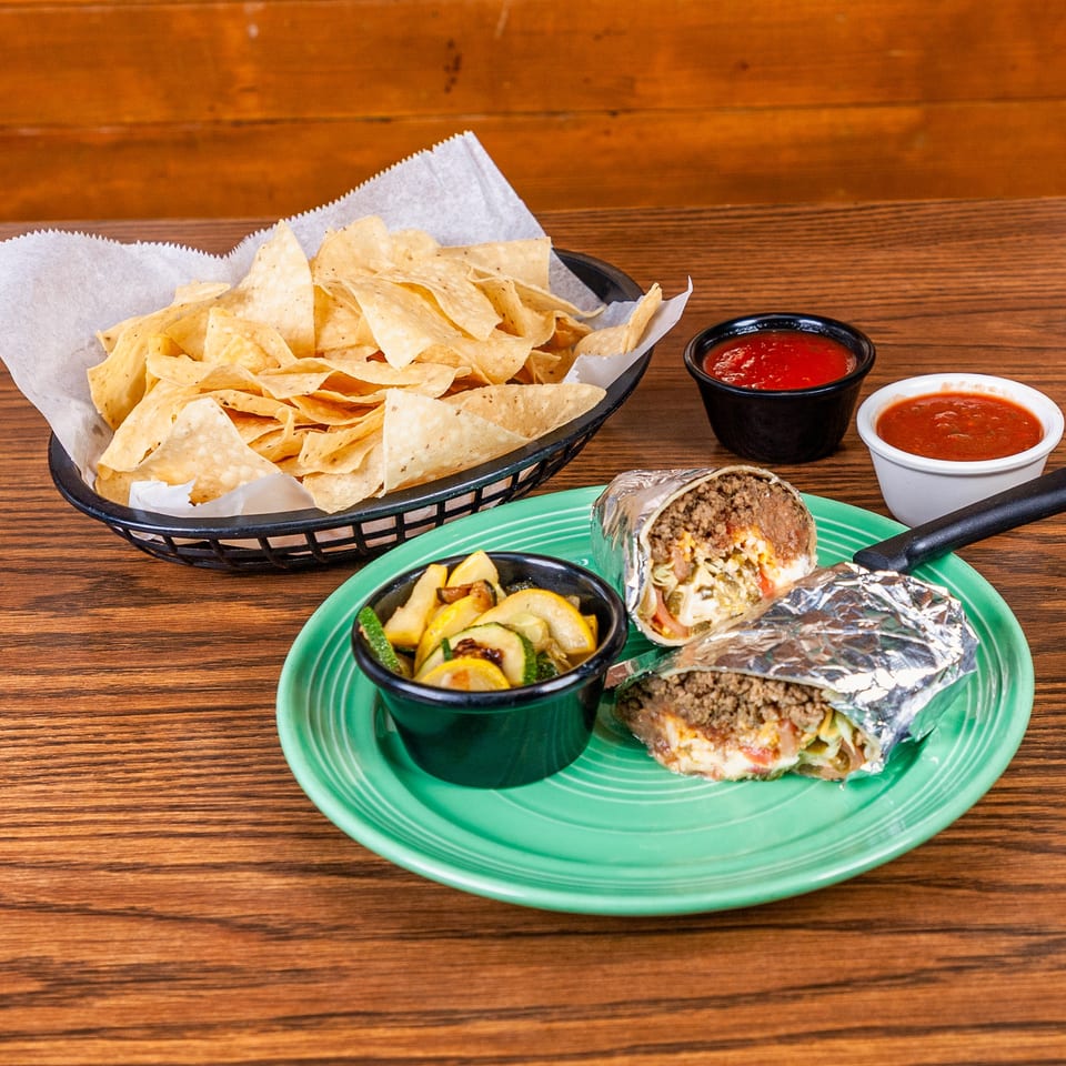 Two burritos, roasted vegetables, tortilla chips, and salsa on a green plate and wooden table.