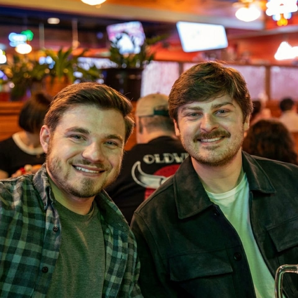 Two smiling men pose for a photo in a dimly lit, busy restaurant with a beer on the table.