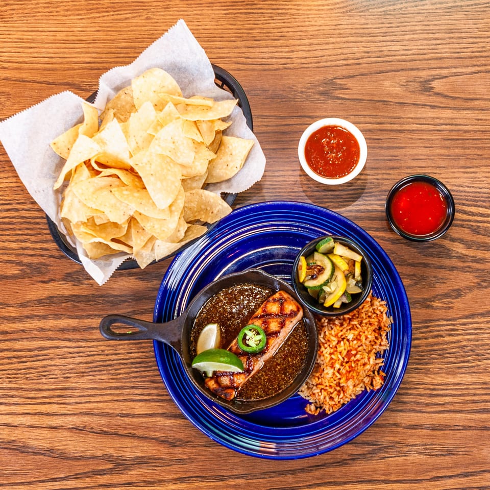 Grilled salmon in a skillet with rice, vegetables, tortilla chips, and salsa on a wooden table.