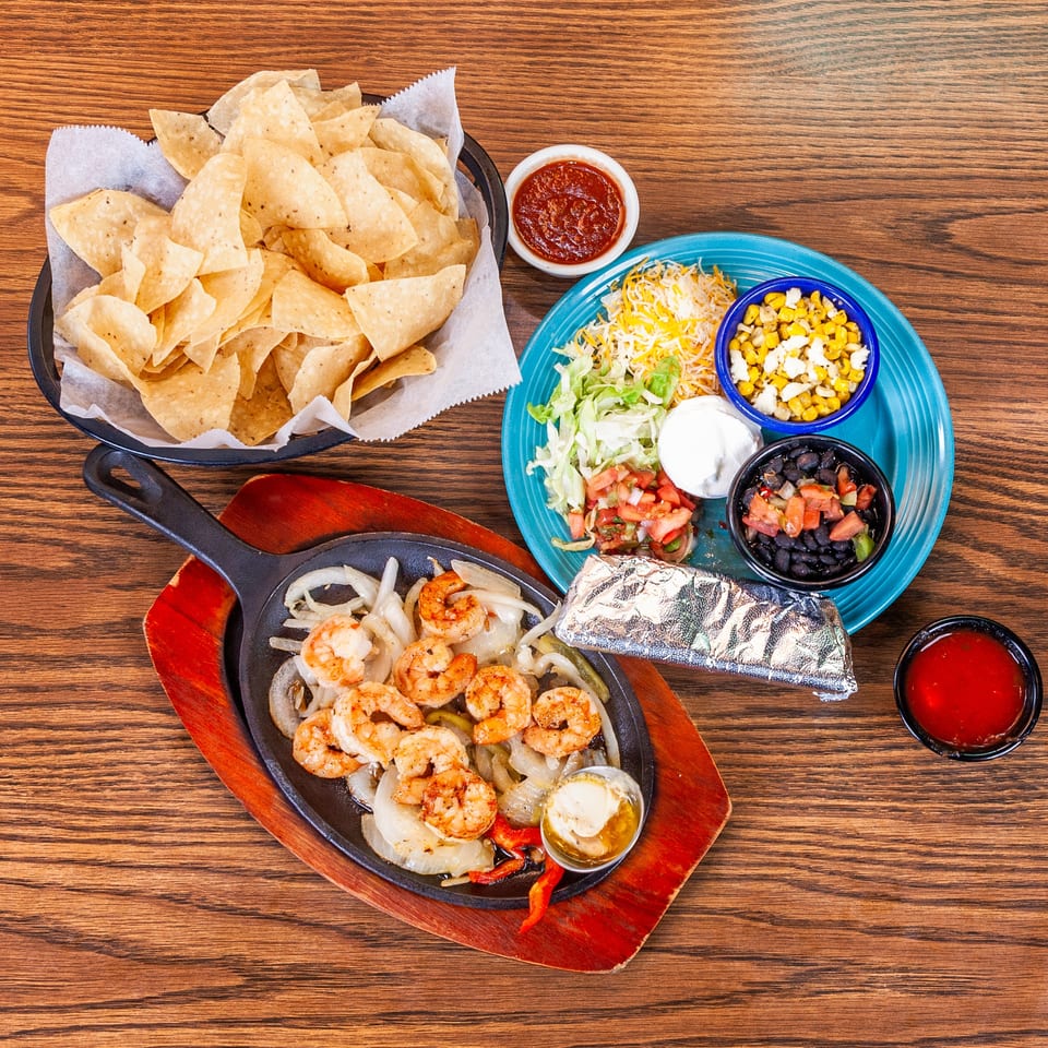 Shrimp fajitas with onions and peppers, a side plate of toppings, tortilla chips, and salsa on a wooden table.
