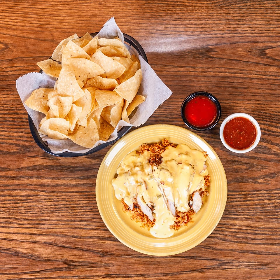 Chicken and rice with cheese sauce, tortilla chips, and two salsas on a wooden table.