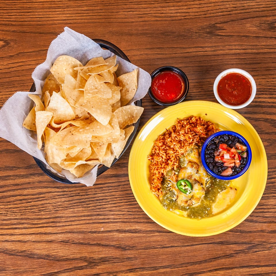 Mexican meal with green enchiladas, rice, black beans, tortilla chips, and salsa on a wooden table.