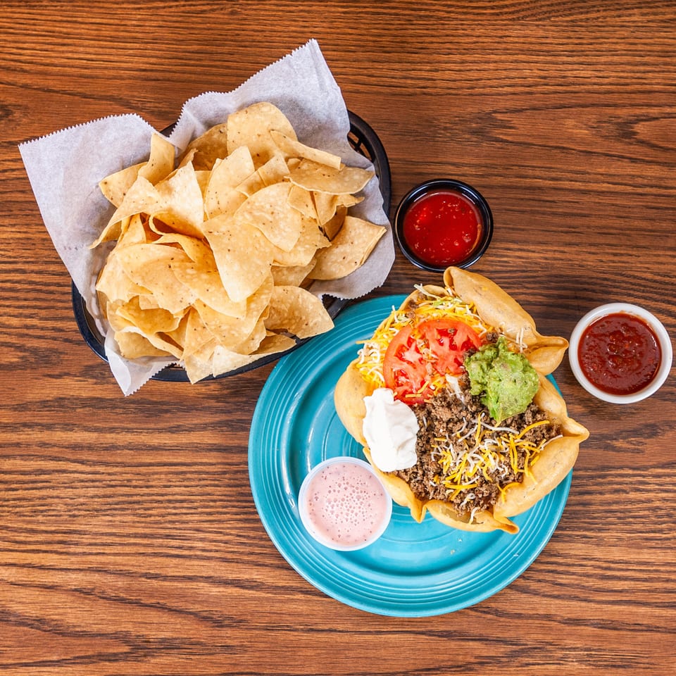 Taco salad with beef, cheese, lettuce, tomato, guacamole, sour cream, chips, and salsa on a wooden table.