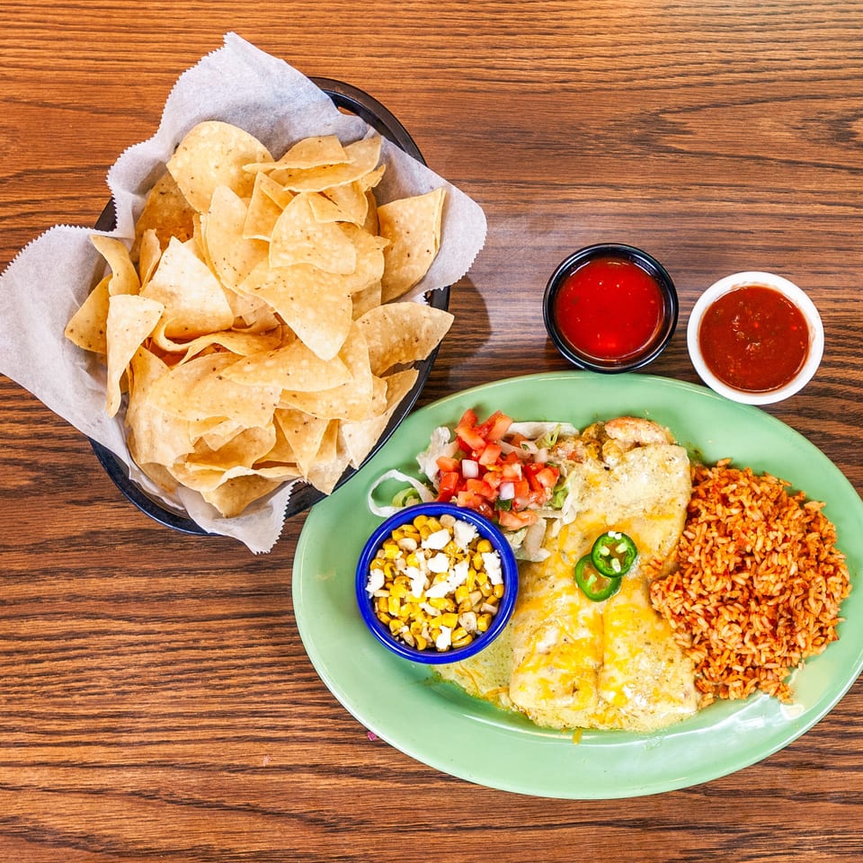 Enchiladas with rice, corn, and pico de gallo on a green plate, alongside a basket of chips and salsa.