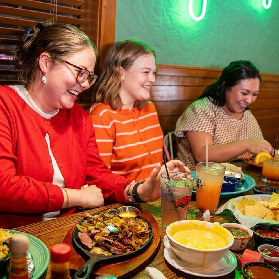 Three women laughing and enjoying a meal with drinks and various dishes at a restaurant.